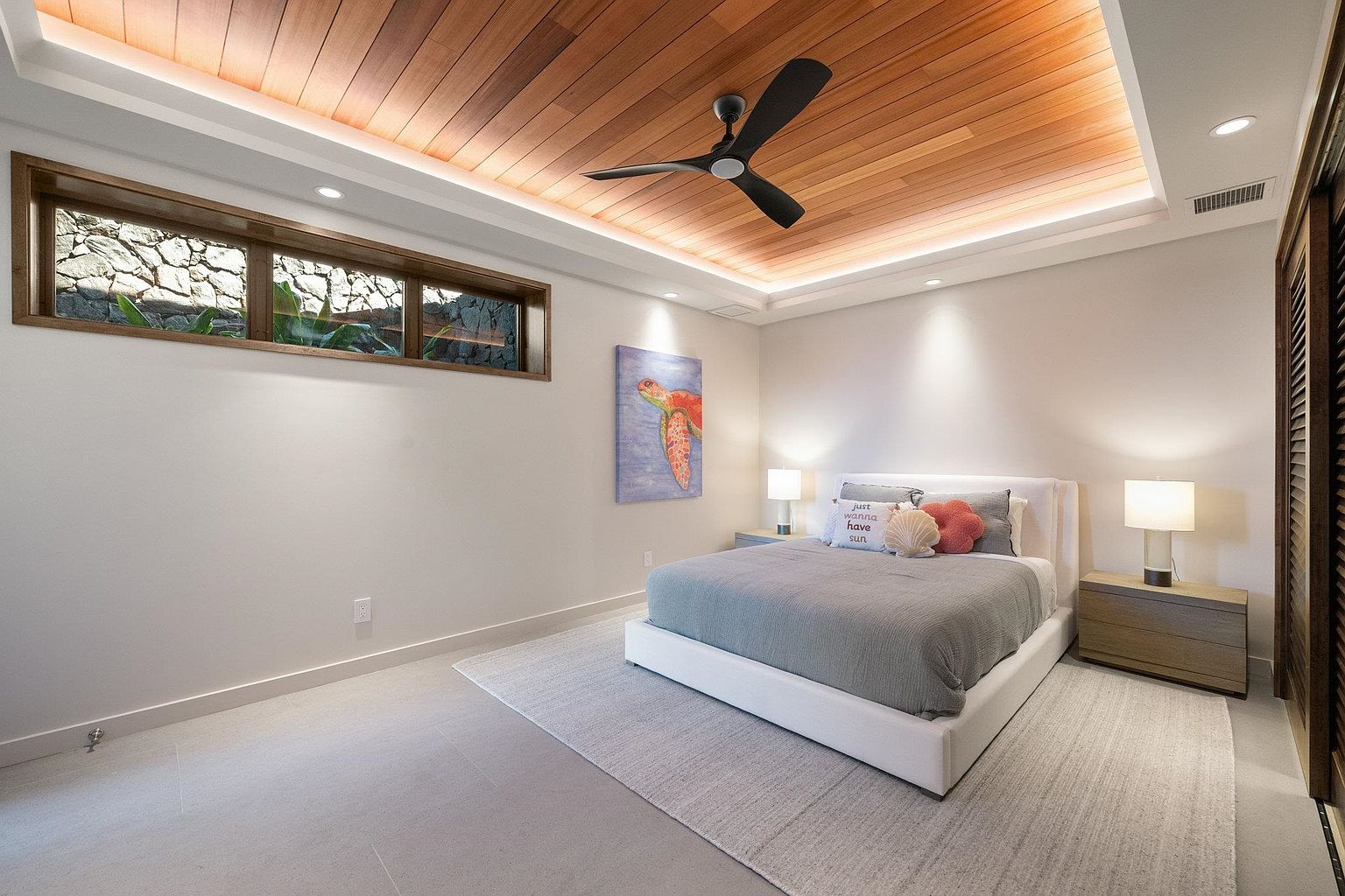This is a primary bedroom featuring a modern design with a wooden plank ceiling and recessed lighting. The room includes a bed with a light gray comforter, two nightstands with lamps, and a piece of artwork on the wall. A long, narrow window provides natural light and a view of the exterior.