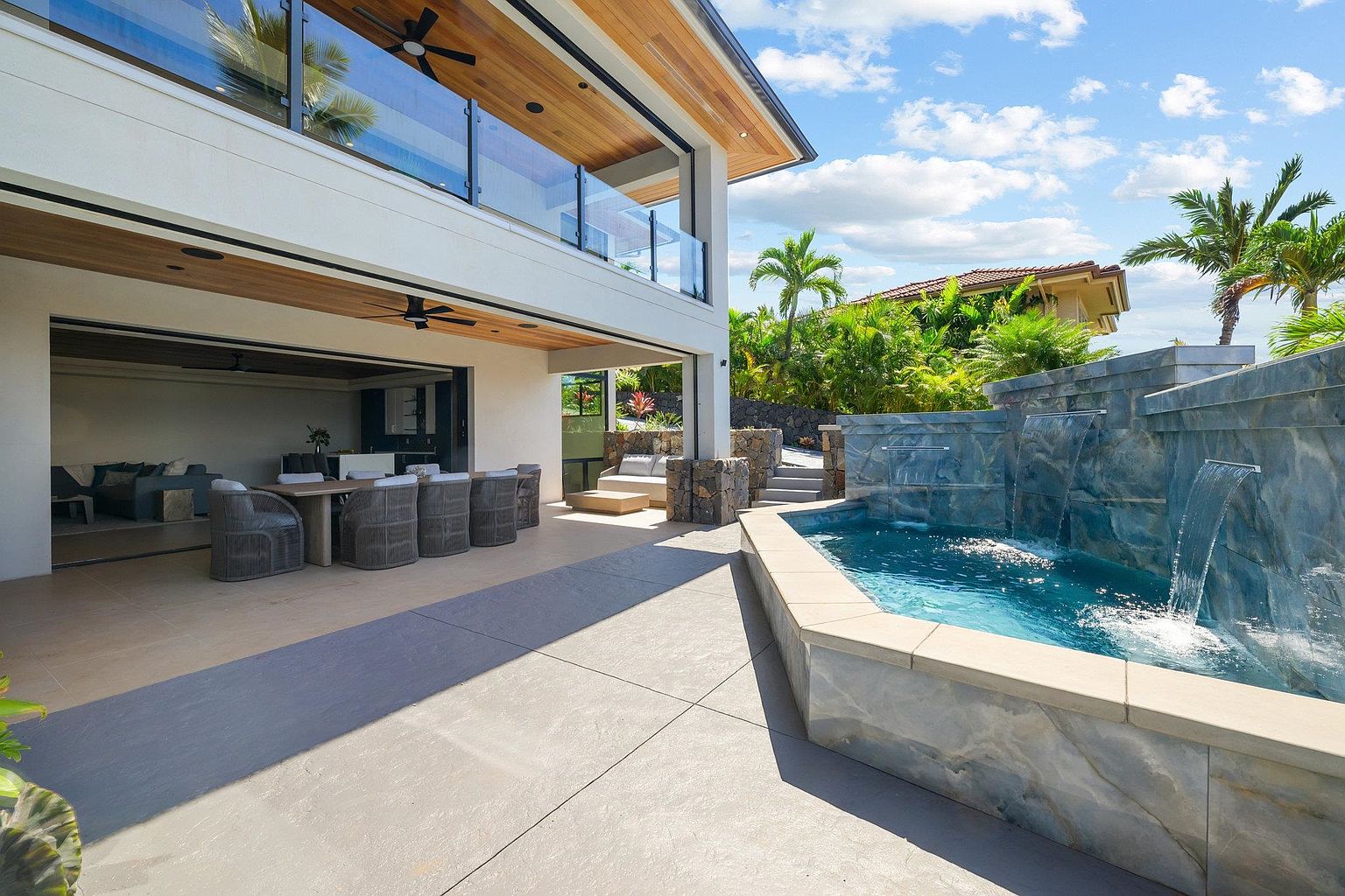 This image showcases a luxurious outdoor patio area with a dining set, comfortable seating, and a stunning water feature with a pool. The patio seamlessly extends from the interior living space, creating an inviting indoor-outdoor flow. The architectural design is modern and elegant, complemented by lush landscaping and a clear blue sky.