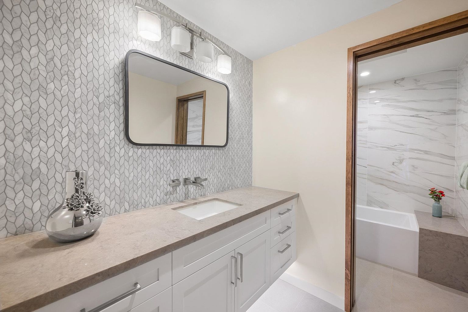 This is a well-lit primary bathroom featuring a long vanity with white cabinets and a light-colored countertop. A decorative silver vase sits on the counter, and a large mirror hangs above the sink, reflecting a glimpse of the adjacent shower/tub area. The walls are adorned with a stylish mosaic tile pattern, adding texture and visual interest to the space.