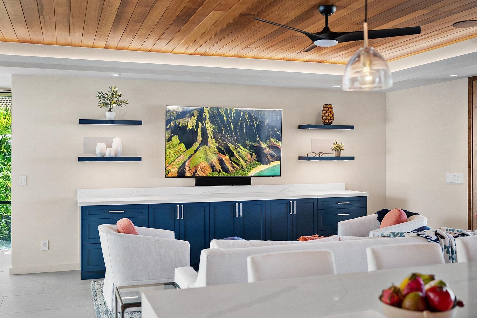 This interior shot showcases a modern living room with a focus on a large flat-screen TV mounted above a blue cabinet with a white countertop. The room features white armchairs and a sofa, complemented by a light wood ceiling and a contemporary pendant light. The overall impression is one of stylish comfort and relaxation.