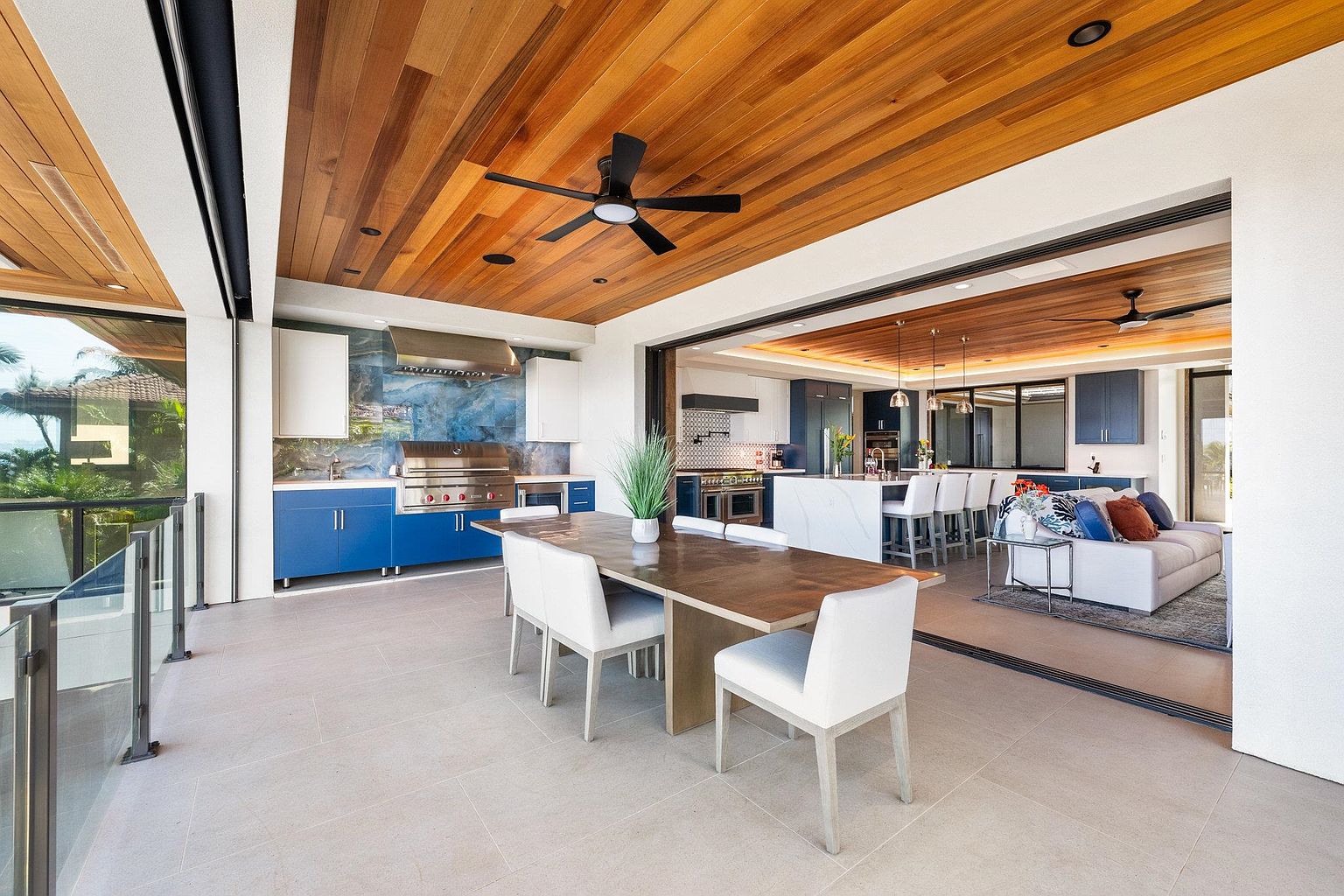 This image showcases a luxurious outdoor patio area seamlessly blending indoor and outdoor living. The space features a dining area with a wooden table and white chairs, an outdoor kitchen with stainless steel appliances and blue cabinetry, and a smooth transition to an interior living space with a sofa and kitchen island. The wooden ceiling adds warmth, while the open design allows for natural light and views.