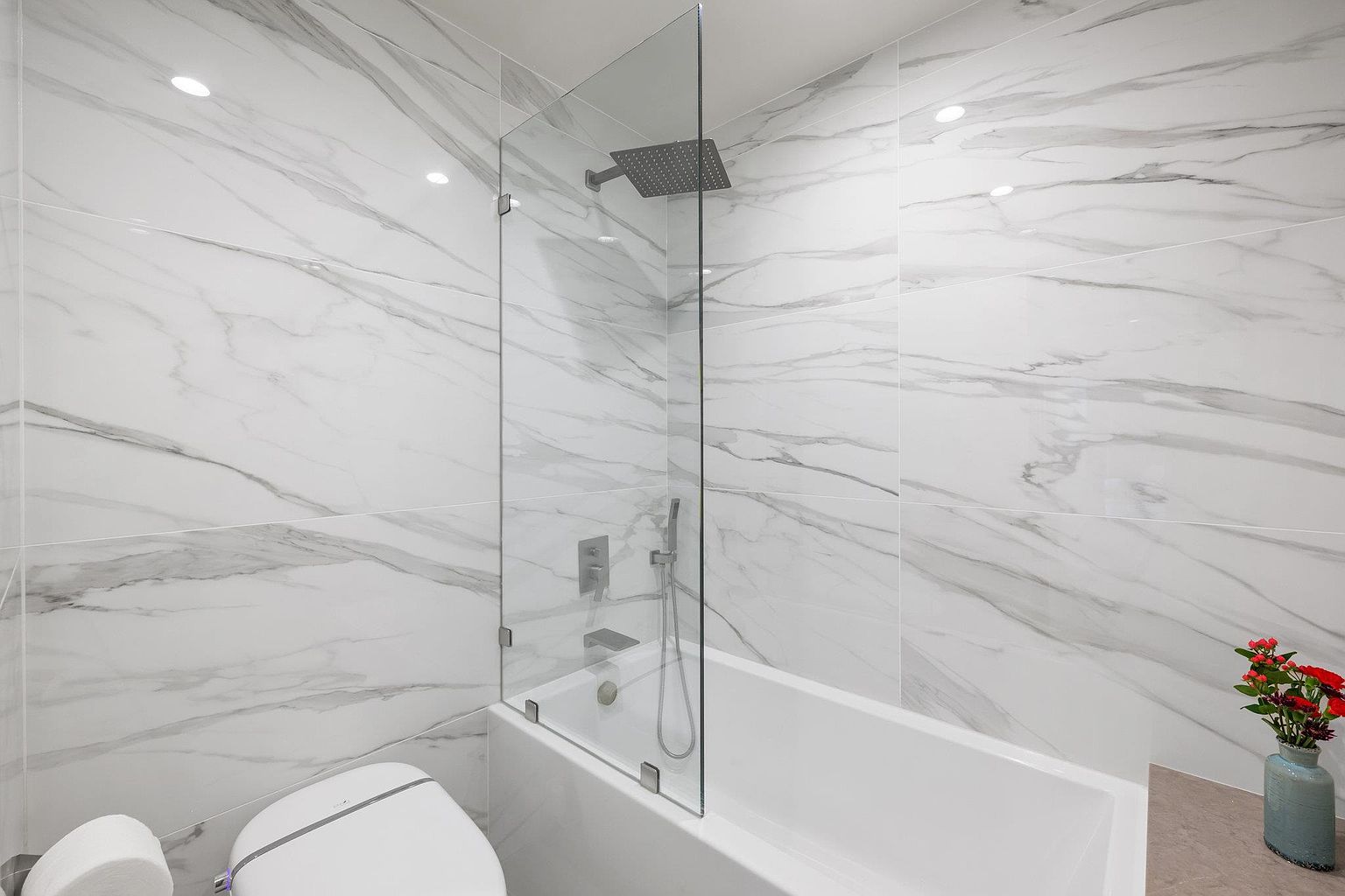 This is a well-lit primary bathroom featuring marble-patterned walls and a modern bathtub with a glass shower screen. A toilet is visible in the foreground, and a vase with red flowers adds a touch of color. The overall impression is clean, bright, and luxurious.