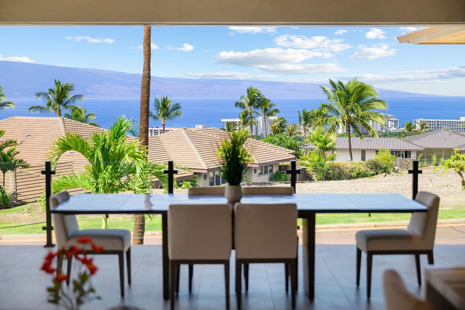 This interior shot showcases a dining room with a stunning ocean view. A dark wood dining table with light-colored chairs sits prominently in the foreground, with a decorative plant centerpiece. The expansive window frames a picturesque scene of palm trees, rooftops, and the ocean under a partly cloudy sky, creating a luxurious and inviting atmosphere.