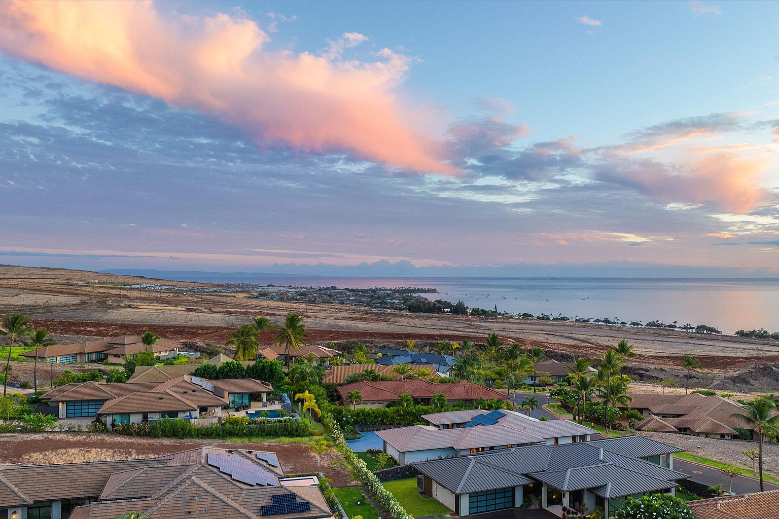 This aerial view showcases a luxury residential neighborhood with well-maintained homes, tropical landscaping, and rooftop solar panels. The scene overlooks a coastal area where the land meets the ocean. The image conveys a sense of upscale living in a desirable location with beautiful ocean views and a serene atmosphere.
