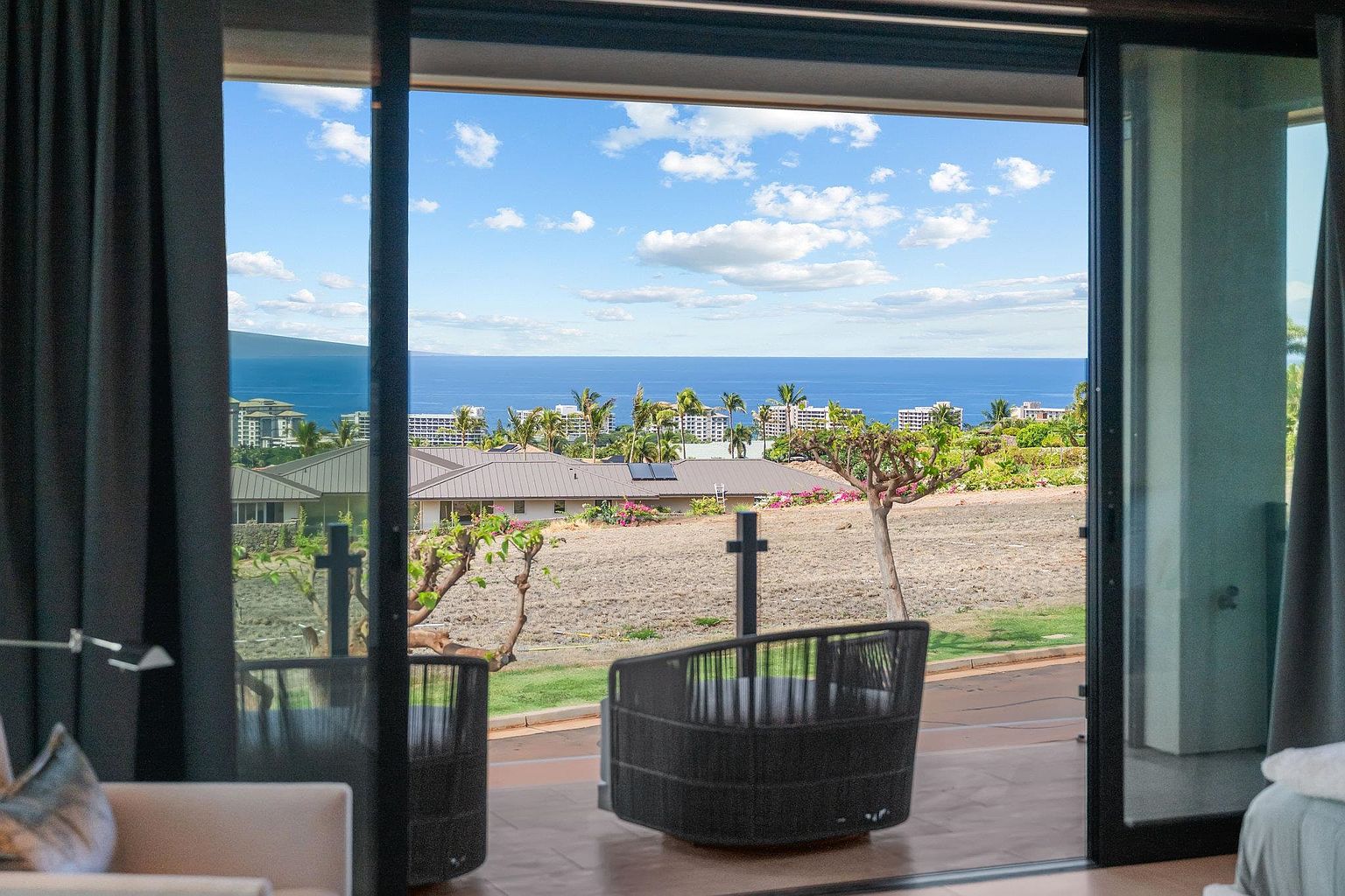 This image showcases a patio or balcony with a stunning ocean view. The outdoor seating area features modern wicker chairs, and the space is framed by sliding glass doors and elegant curtains. The view includes lush greenery, residential buildings, and the vast expanse of the ocean under a partly cloudy sky, creating a serene and luxurious atmosphere.