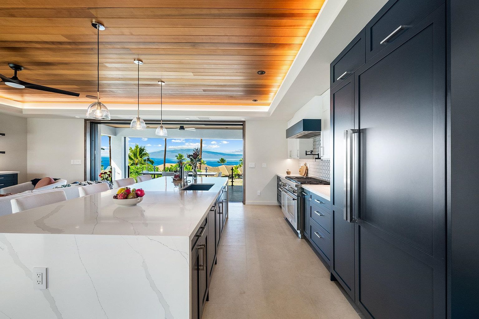 This is a bright, modern kitchen featuring a large island with a white countertop and dark cabinetry. The kitchen is open to the outside with sliding glass doors that offer a stunning ocean view. High-end appliances and a wood-paneled ceiling add to the luxurious feel of the space.