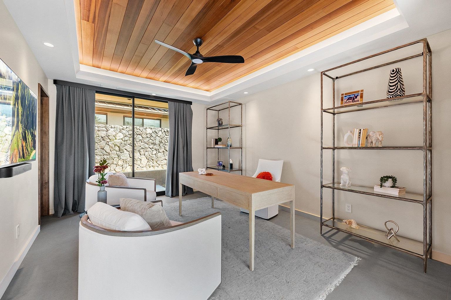 This is an interior shot of a modern office space featuring a wooden ceiling with recessed lighting and a ceiling fan. The room includes a light wood desk, two white armchairs, and two metal shelving units displaying decorative items. A large window with gray curtains provides natural light and a view of a stone wall, creating a serene and stylish workspace.