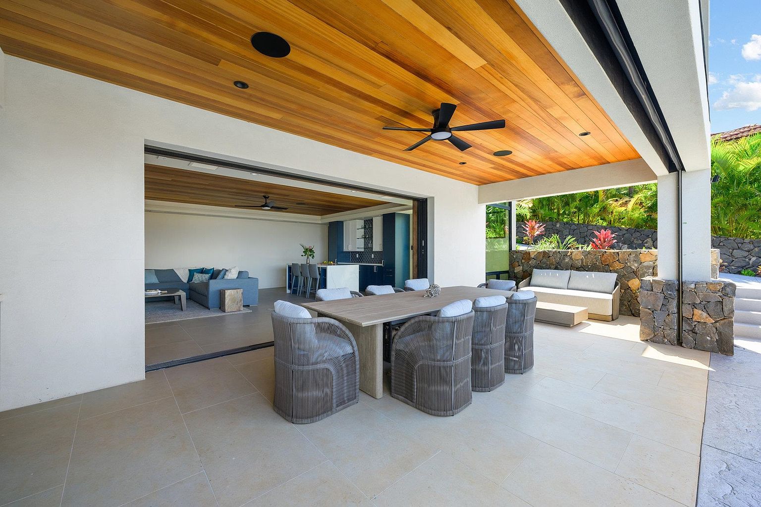 This image showcases a luxurious outdoor patio area, seamlessly blending indoor and outdoor living. The space features a dining set with woven chairs, a comfortable lounge area, and a wooden ceiling with a ceiling fan. Large sliding glass doors open to an interior living space, while a stone wall and lush greenery provide privacy and a natural backdrop.