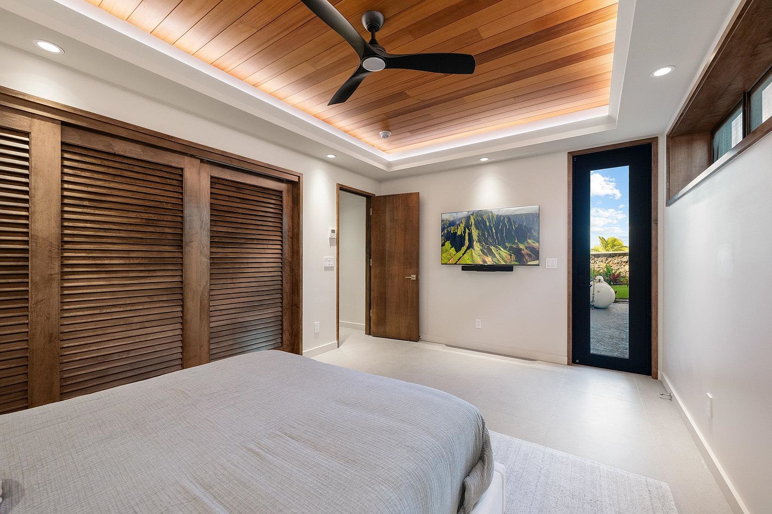 This is a primary bedroom featuring a wooden plank ceiling with recessed lighting and a ceiling fan. The room has a large bed, a wall-mounted television, and a sliding door closet. A door leads to another room, and a glass door provides access to an outdoor area, creating a bright and airy atmosphere.