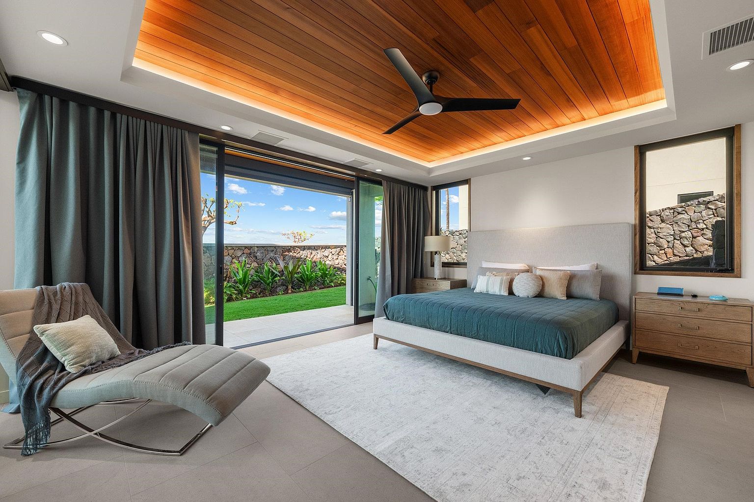 This is a luxurious primary bedroom featuring a king-size bed with a teal comforter and decorative pillows. The room has a wood-paneled ceiling with recessed lighting and a modern ceiling fan. Sliding glass doors open to a patio and garden, providing ample natural light and a seamless indoor-outdoor flow, while a chaise lounge offers a comfortable seating area.