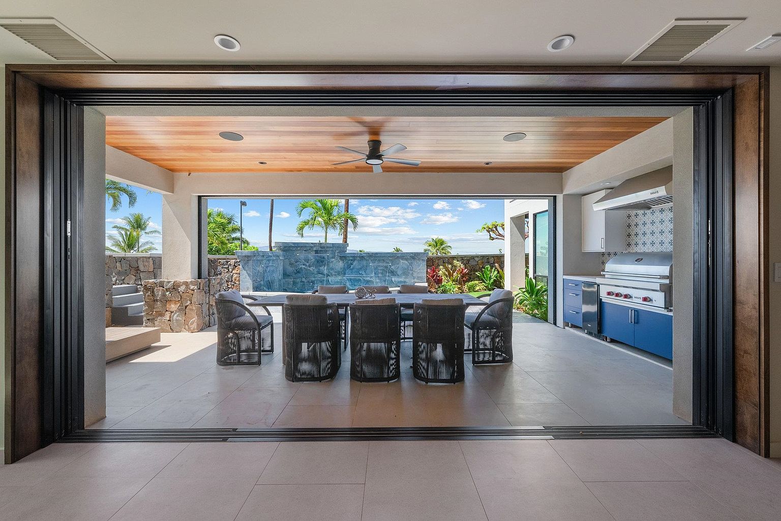 This image showcases a luxurious outdoor patio area, seamlessly blending indoor and outdoor living. The space features a large dining table with seating for eight, an outdoor kitchen with stainless steel appliances, and a stunning view of tropical landscaping and a water feature. The open-air design, coupled with the high-end finishes, creates an inviting and sophisticated atmosphere perfect for entertaining.