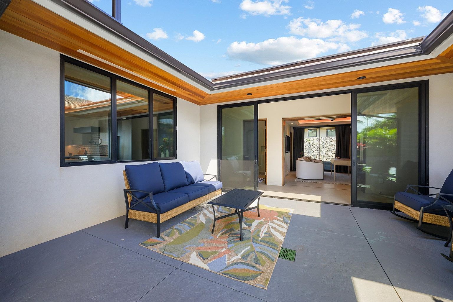 This image showcases a stylish outdoor patio area, featuring a comfortable blue sofa and a modern coffee table set on a colorful rug. The patio is seamlessly connected to the interior through large sliding glass doors, offering an inviting indoor-outdoor living experience. The space is well-lit and appears to be part of a contemporary home design.