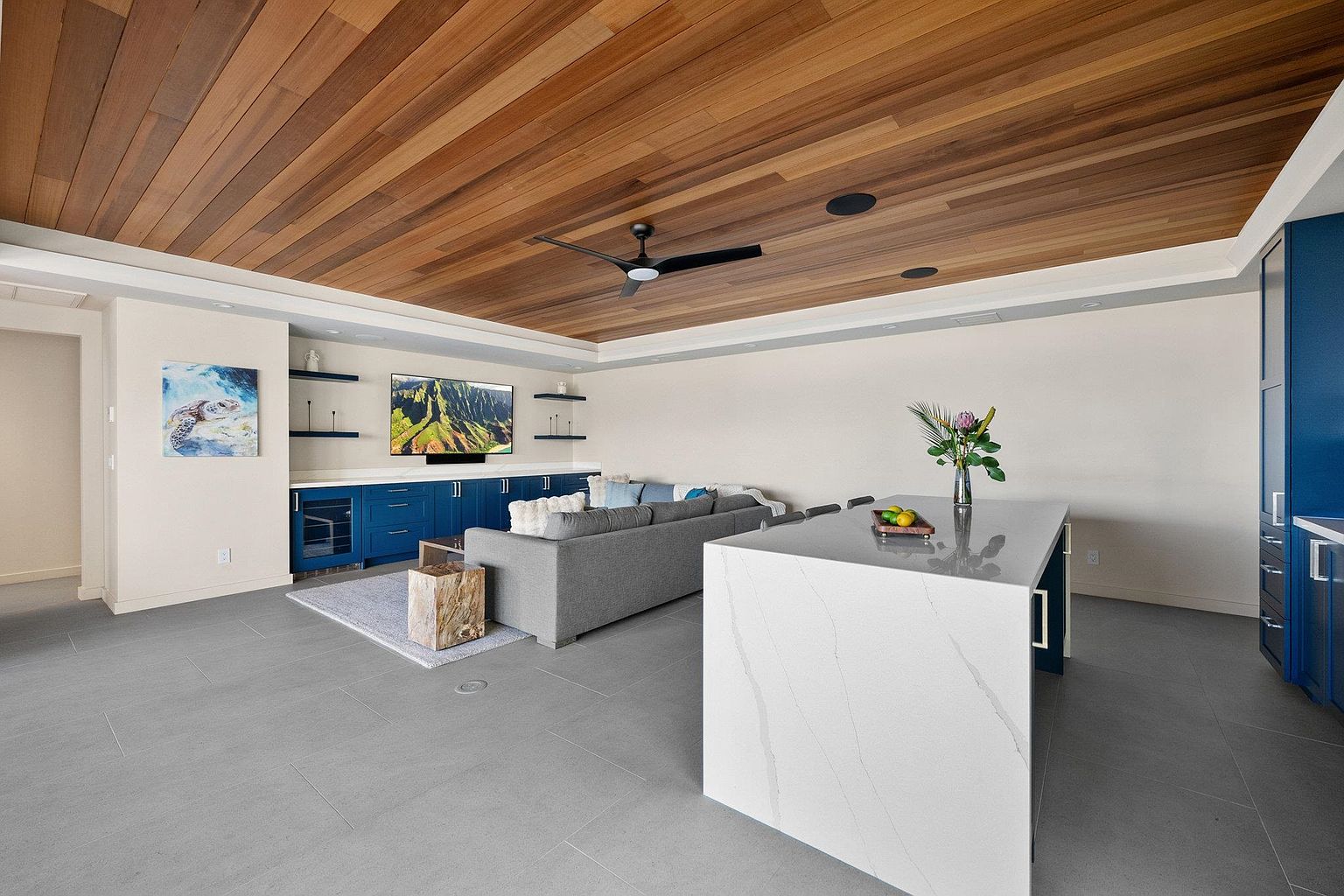 This interior shot showcases a modern living room with a unique wooden plank ceiling and recessed lighting. The room features a gray sectional sofa, a blue built-in entertainment center with shelving, and a large kitchen island with a white countertop. The flooring is a sleek gray tile, and the overall aesthetic is contemporary and inviting.