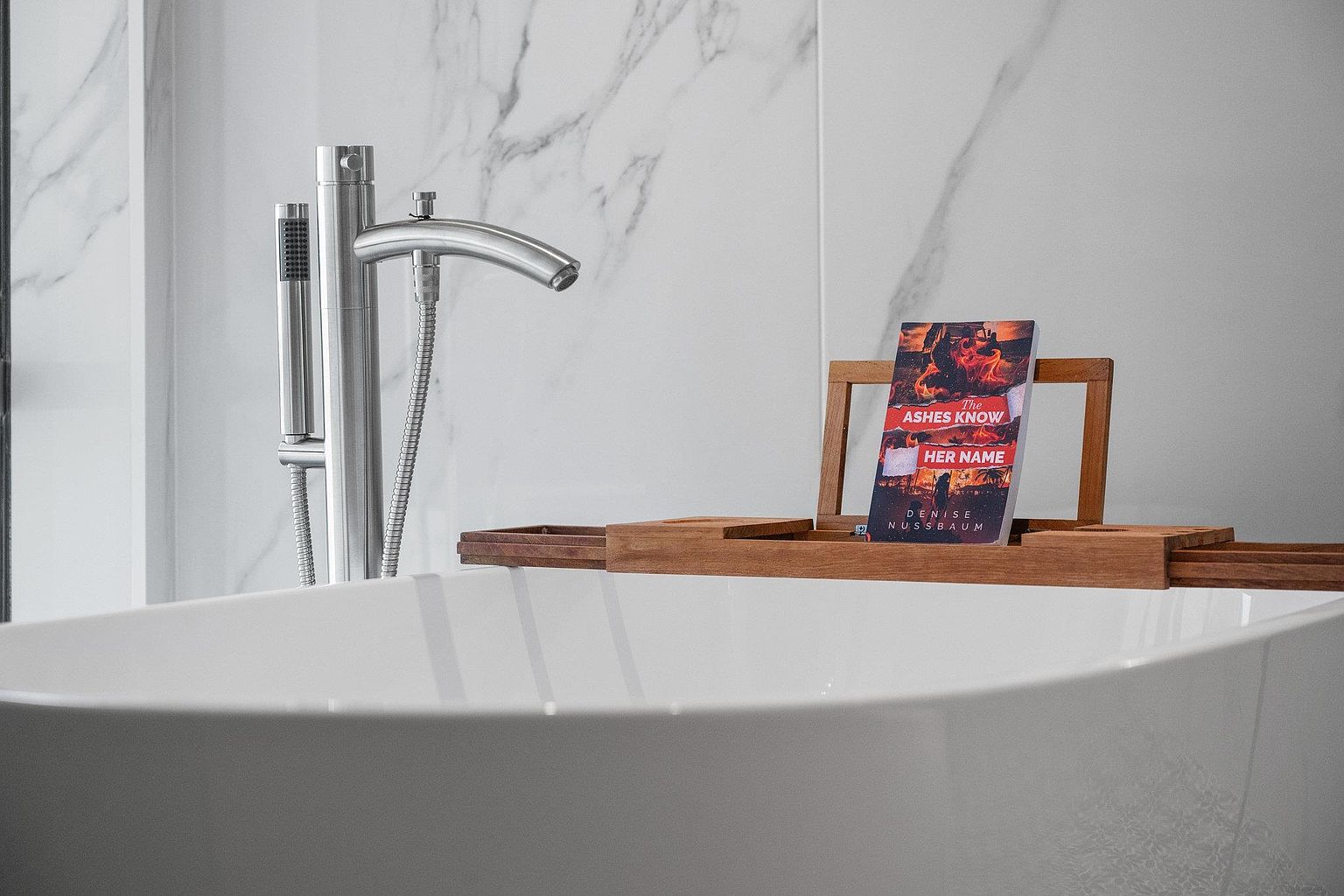 This is a close-up shot of a modern primary bathroom featuring a freestanding bathtub with a sleek, chrome faucet and handheld shower attachment. A wooden bath caddy rests across the tub, holding a book titled "The Ashes Know Her Name." The background showcases a marble-patterned wall, adding a touch of luxury to the scene.