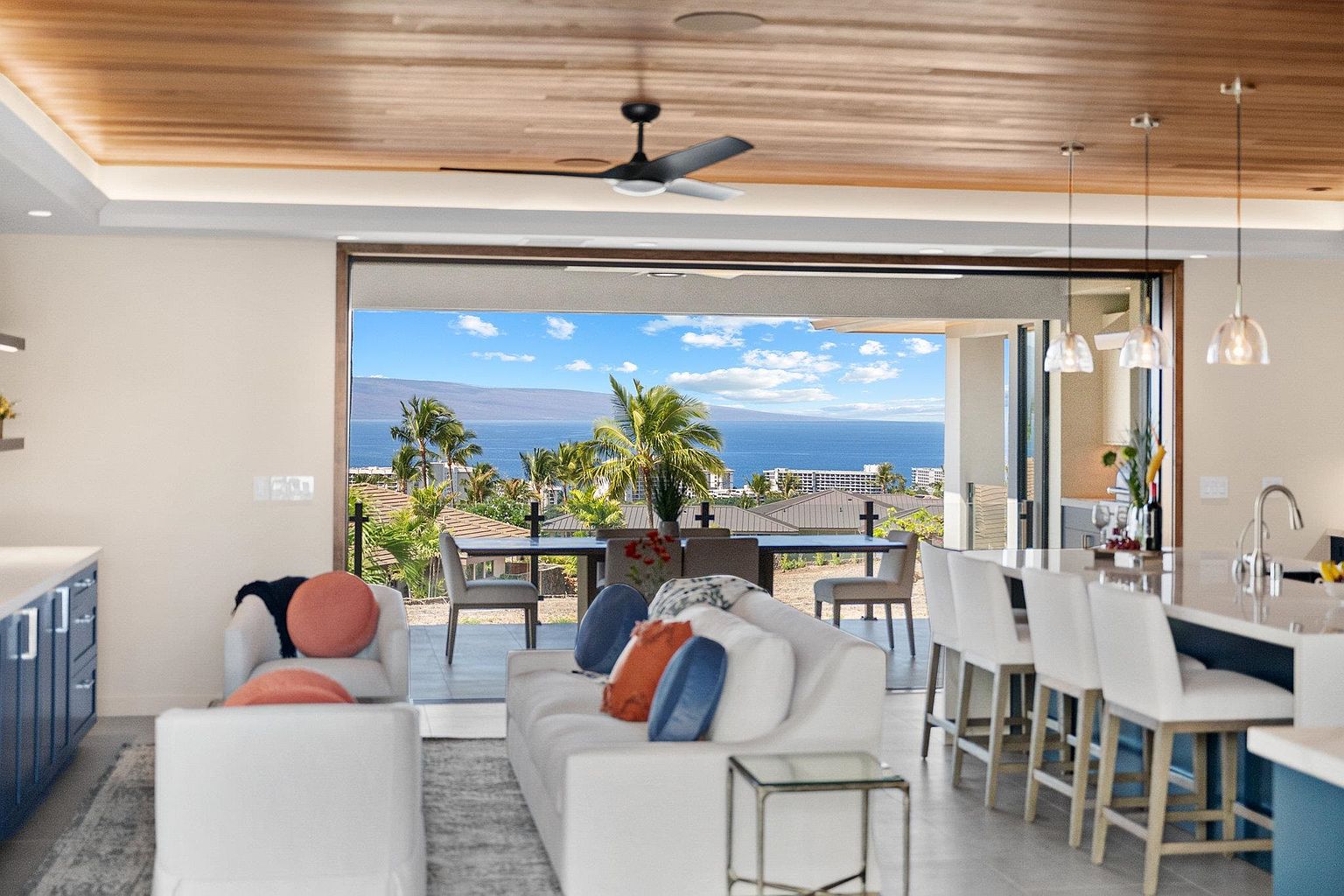 This interior shot showcases a luxurious living room that seamlessly blends indoor and outdoor living. A large sliding glass door opens to a stunning ocean view, with a dining area visible on the patio. The living room features white sofas with colorful pillows, a modern coffee table, and a glimpse of a kitchen island with bar stools, creating an inviting and sophisticated atmosphere.