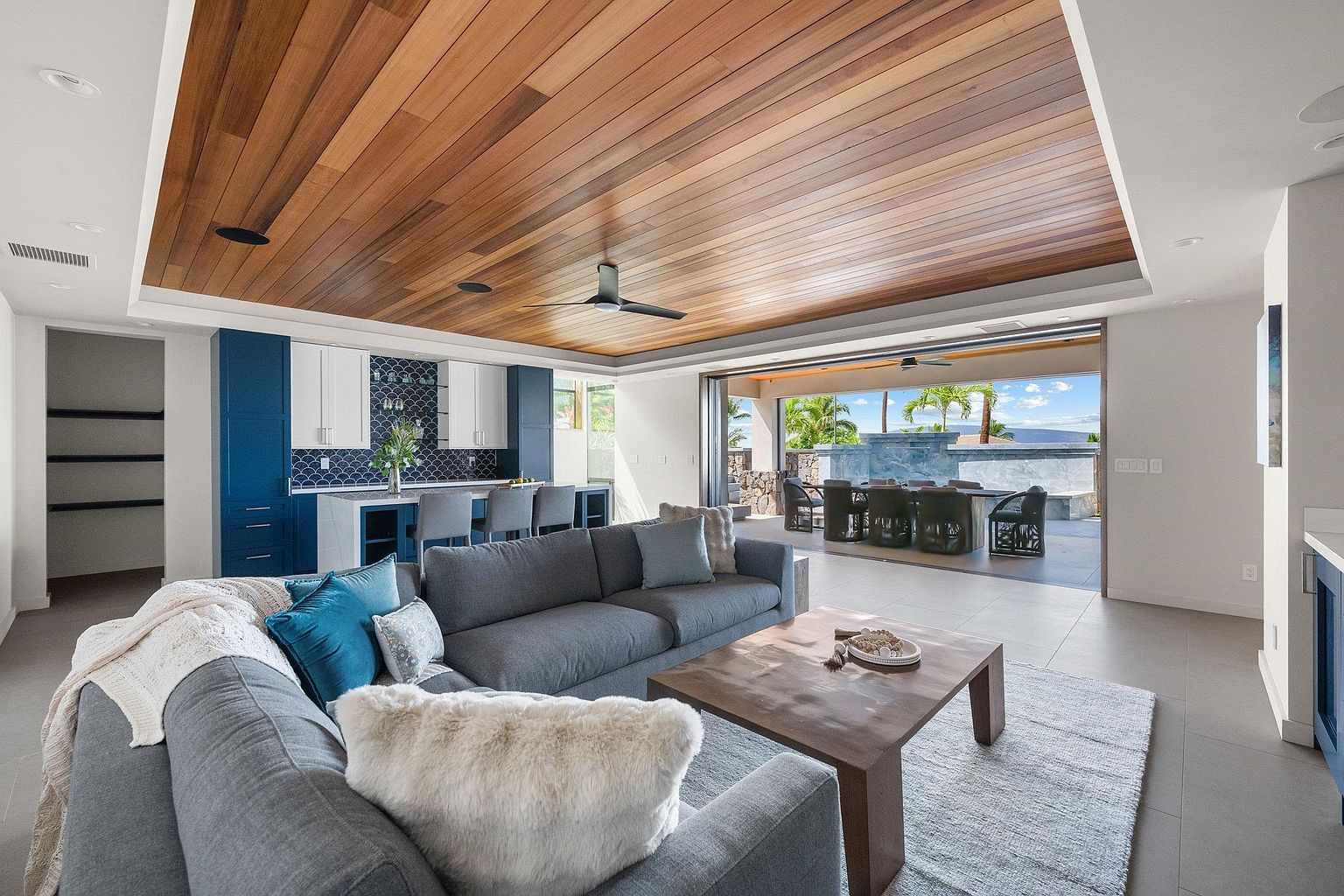 This is an interior shot of a modern living room that seamlessly blends indoor and outdoor living. The room features a large gray sectional sofa with decorative pillows, a wooden coffee table, and a light-colored area rug. A wooden plank ceiling adds warmth, while a retractable glass wall opens to an outdoor dining area and pool, creating an airy and inviting space.