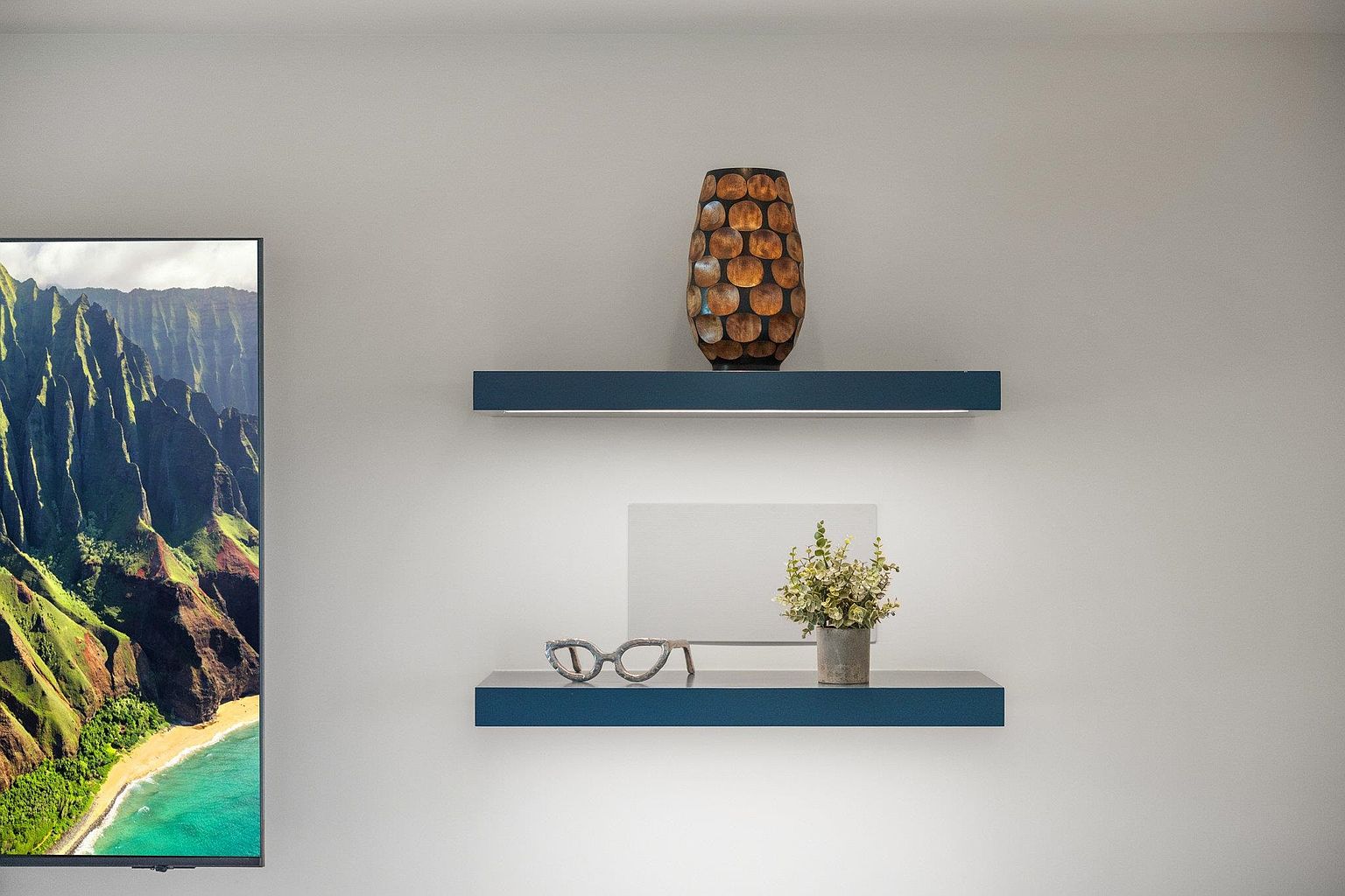The image showcases a modern living room detail with two floating shelves mounted on a light gray wall. The top shelf features a decorative brown vase, while the lower shelf holds a pair of glasses, a small potted plant, and a white picture frame. To the left, a television displays a scenic landscape, adding a touch of nature to the interior.