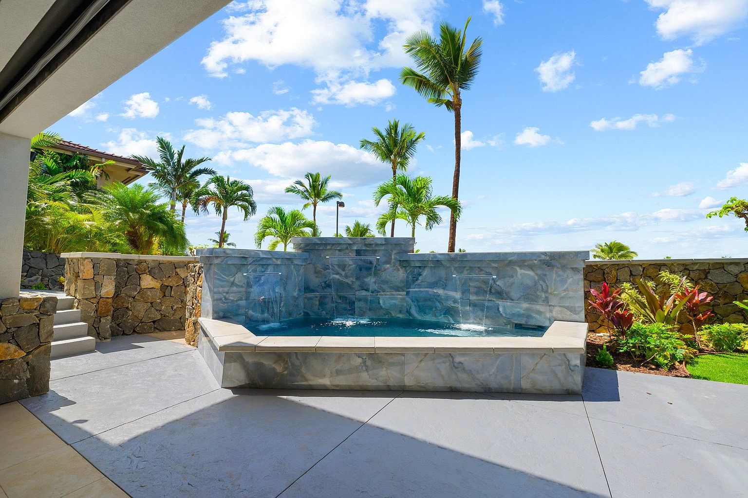 This image showcases a luxurious outdoor spa area with a multi-tiered stone waterfall feature. Palm trees dot the landscape under a bright blue sky with scattered clouds, creating a tropical ambiance. The spa is surrounded by a well-maintained patio and lush greenery, suggesting a private and serene retreat.