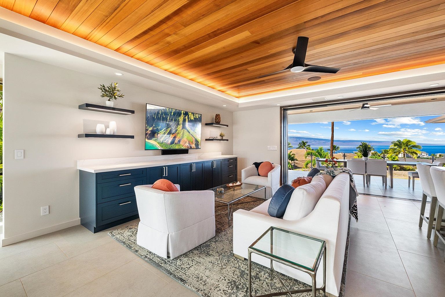 This is a bright and airy living room featuring a wood-paneled ceiling with recessed lighting, a built-in media center with dark blue cabinetry, and comfortable white seating. Large sliding glass doors open to an outdoor dining area with ocean views, creating a seamless indoor-outdoor living experience. The room is decorated in a modern style with a neutral color palette and glass tables.