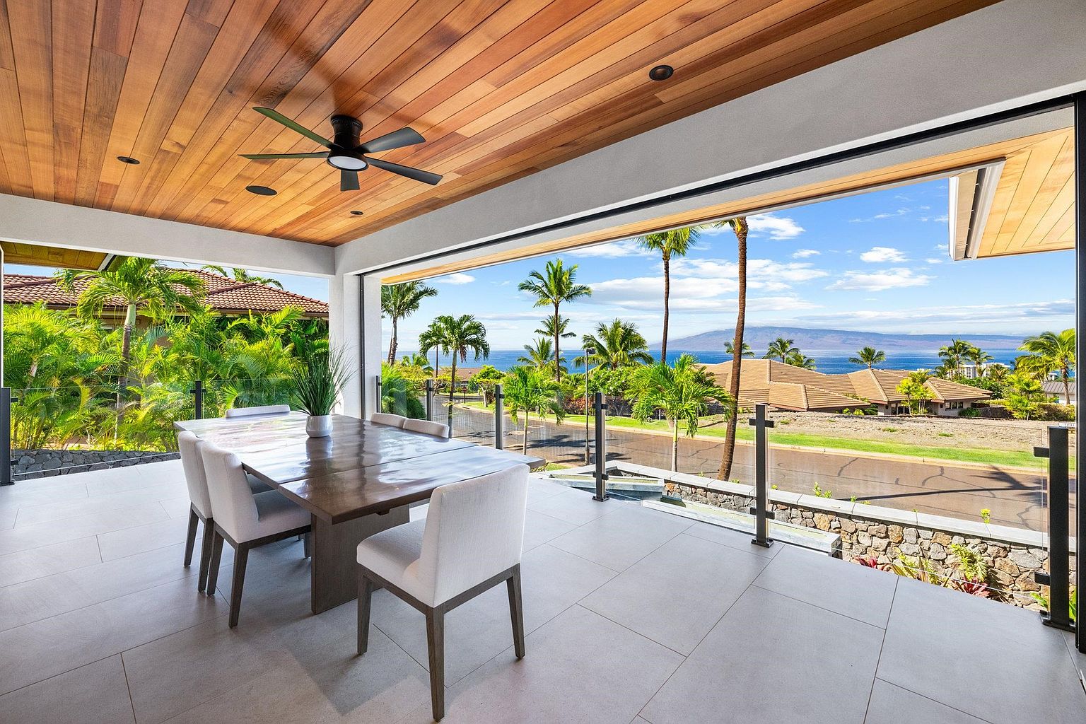This image showcases a luxurious outdoor dining area on a patio or balcony. The space features a wooden ceiling with a ceiling fan, a large dining table with white chairs, and an expansive view of the ocean and palm trees. The open design seamlessly blends indoor and outdoor living, creating an inviting and elegant atmosphere.