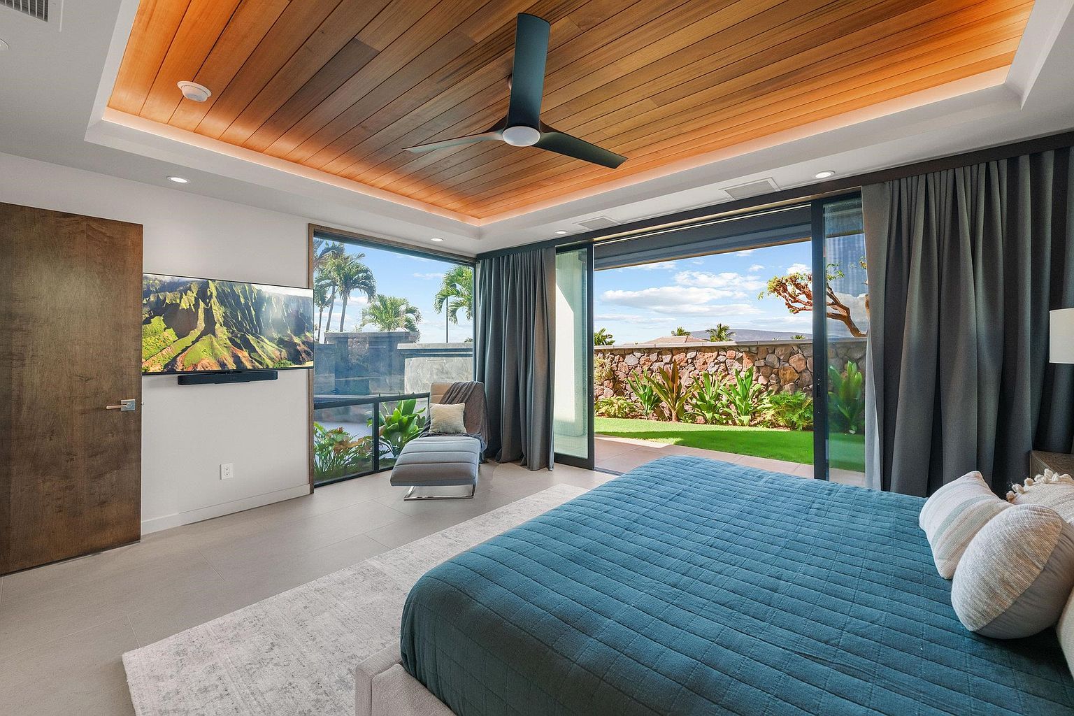 This is a luxurious primary bedroom featuring a king-size bed with a blue quilted cover, complemented by neutral-toned pillows. The room boasts a wooden plank ceiling with recessed lighting and a modern ceiling fan. Large sliding glass doors open to a well-manicured lawn and stone wall, creating a seamless indoor-outdoor living experience. A chaise lounge sits near a window with views of palm trees.