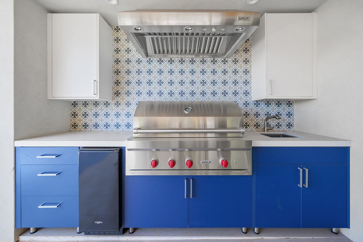 This image showcases an outdoor kitchen area, featuring a stainless steel grill with red knobs, blue cabinetry, and a patterned backsplash. The countertops are light-colored, and there's a built-in beverage cooler. The overall design is modern and functional, perfect for outdoor entertaining.