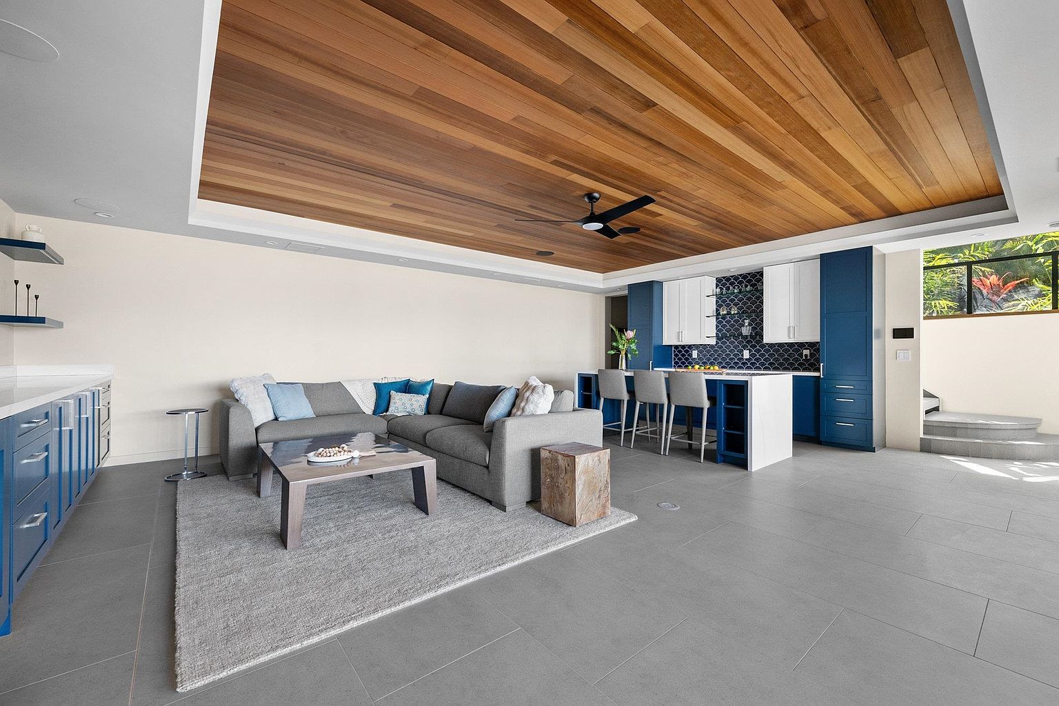 This interior shot showcases a modern living room and kitchen area with a warm, inviting atmosphere. The living room features a gray sectional sofa, a light gray rug, and a wooden coffee table, while the adjacent kitchen boasts blue and white cabinetry with a unique backsplash. The wood-paneled ceiling adds a touch of elegance and warmth to the space.