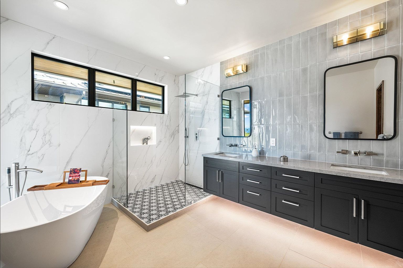 This is a luxurious primary bathroom featuring a freestanding soaking tub with a wooden tray, a glass-enclosed shower with patterned tile flooring, and a double vanity with dark cabinetry and light countertops. The walls are adorned with a combination of marble and light gray tiles, complemented by modern lighting fixtures and framed mirrors. The overall impression is clean, modern, and spa-like.