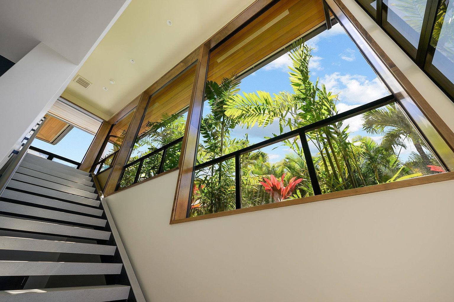 This interior shot showcases a modern hallway with a staircase featuring white and black steps. Large, angled windows provide a view of lush tropical foliage, creating a connection with the outdoors. The design emphasizes natural light and a clean, contemporary aesthetic.