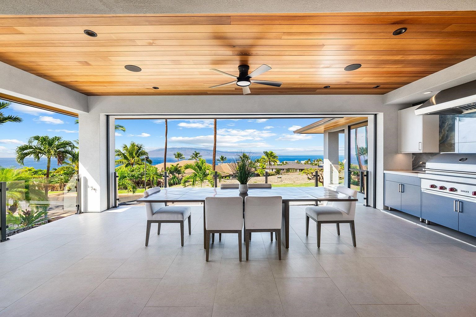 This interior shot showcases a dining area with a stunning view. The dining table is set with chairs, and large sliding glass doors open to a tropical landscape with palm trees and ocean views. The ceiling is wood paneled, and the flooring is light tile, creating a bright and airy atmosphere.