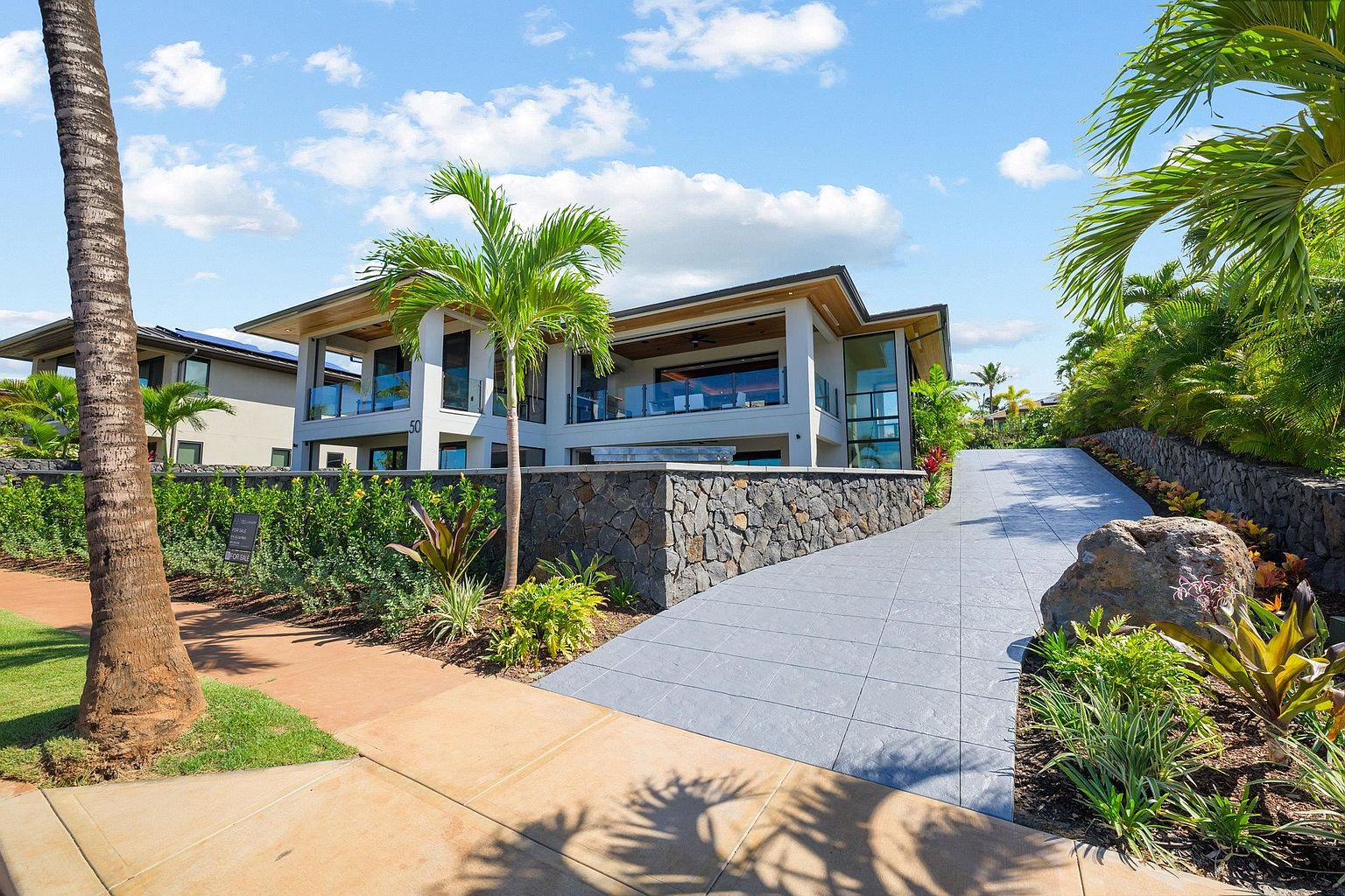 This is a front exterior view of a modern, two-story home with a flat roof and a stone retaining wall. The house features large windows and balconies, surrounded by lush tropical landscaping including palm trees and manicured hedges. A paved driveway leads up to the house, enhancing its curb appeal.