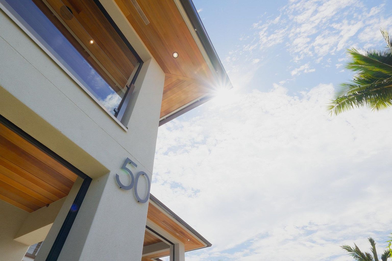 This is a low-angle shot of a modern home's exterior, emphasizing its architectural details. The facade features a light-colored stucco finish, complemented by warm wood accents on the eaves and soffits. The house number '50' is prominently displayed, and a glimpse of the sky with scattered clouds and a palm tree adds a tropical touch.