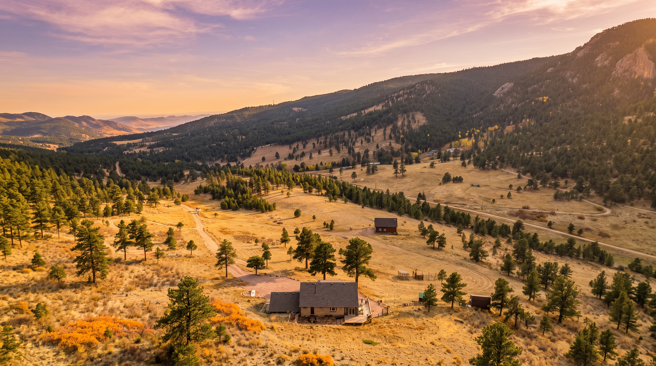 This high-angle aerial view captures a secluded residential property nestled within a vast, rolling landscape of dry, golden grasses and scattered pine trees. The main house features a multi-gabled roofline and a rear deck, positioned prominently in the foreground with a secondary structure visible further up the hillside. The expansive terrain stretches toward a distant mountain range under a bright, clear sky, emphasizing the property's privacy and connection to nature.