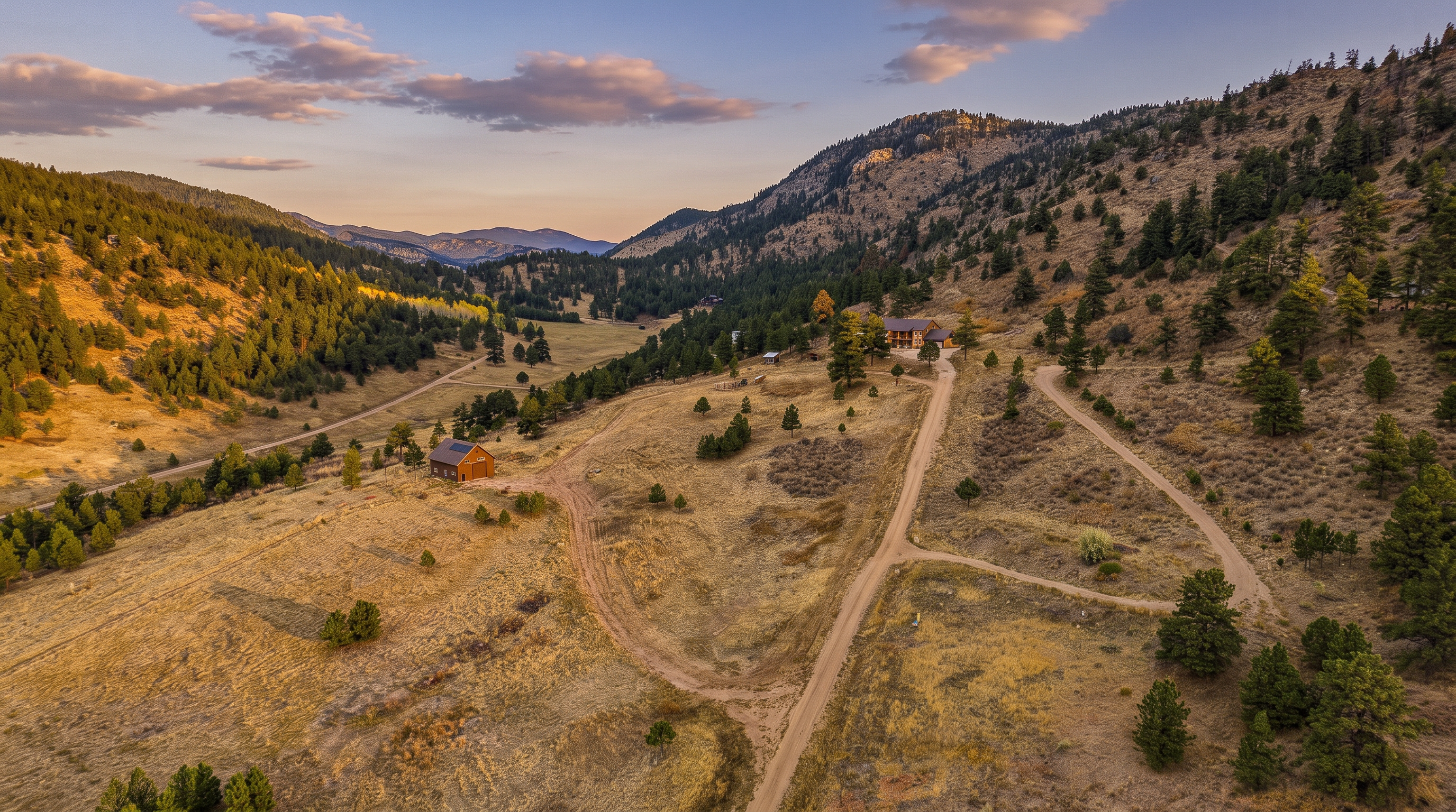 This high-angle aerial view captures a sprawling, rural mountain property set within a vast, golden-hued landscape. The scene features a primary residence nestled on a hillside, a separate detached barn or garage structure, and a network of winding dirt roads connecting the buildings. The surrounding terrain is characterized by rolling hills, scattered evergreen trees, and a distant mountain range under a clear blue sky.