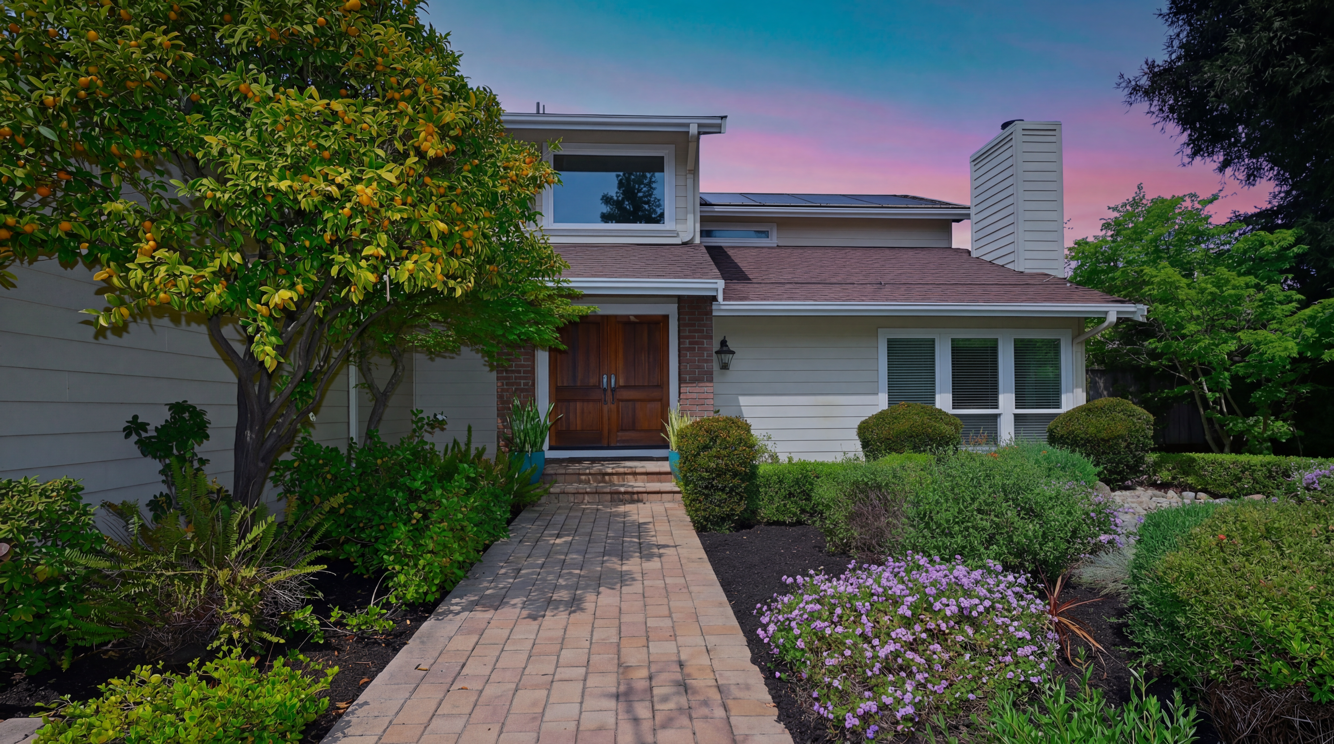 This charming two-story home features a welcoming entryway with classic double wooden doors and a paved walkway leading to the house. The front yard is nicely landscaped with a variety of bushes, purple flowers, and a prominent orange tree, creating excellent curb appeal. The exterior combines traditional siding and brick accents under a beautifully sunset-lit sky.