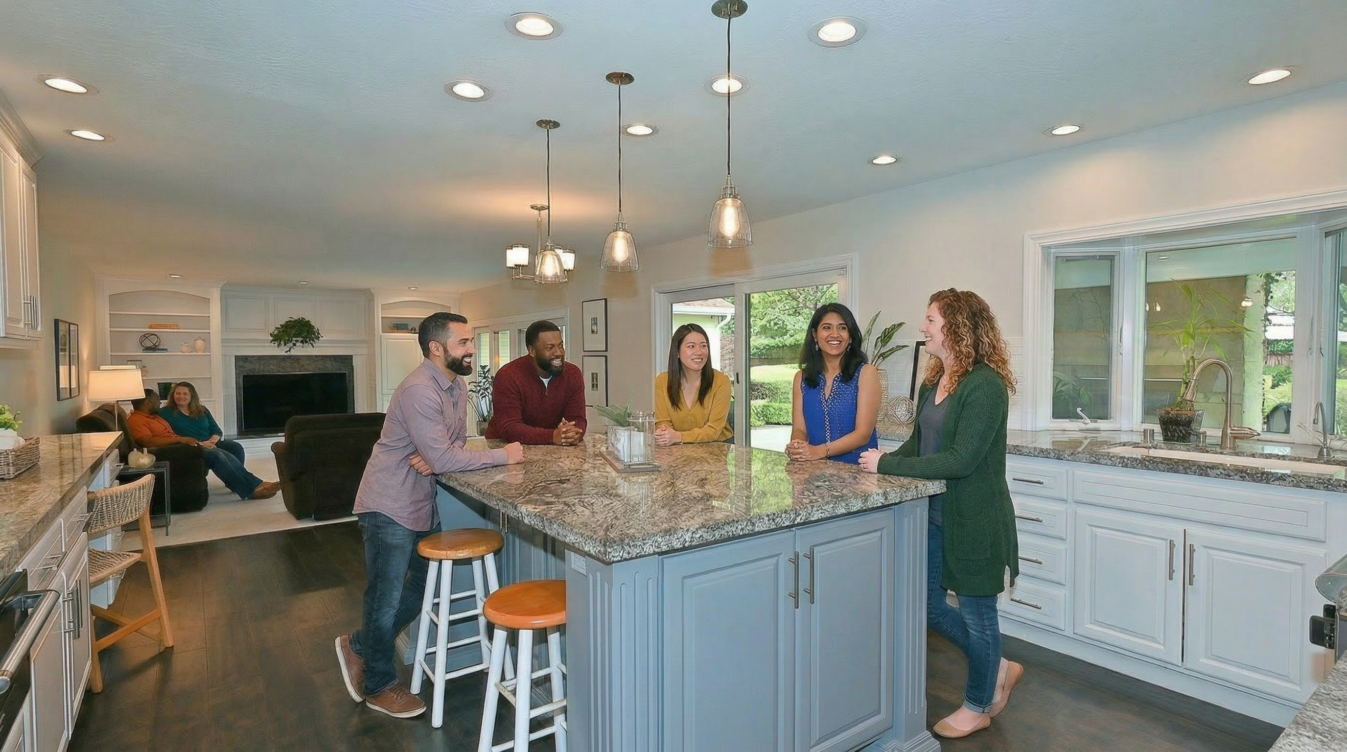This bright, open-concept kitchen features a large central island with a granite countertop, serving as a social focal point for the group of people gathered around it. The space includes white cabinetry, hardwood floors, and a seamless transition into an adjacent living area with a fireplace. The design offers an inviting, contemporary aesthetic, highlighted by modern pendant lighting and high-end finishes suitable for family living.