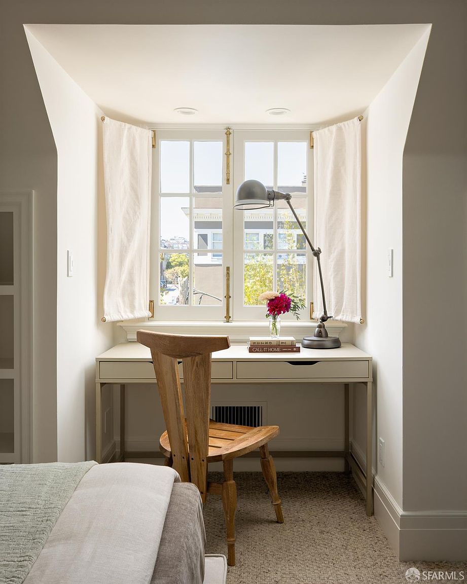 This cozy home office nook is nestled within a windowed alcove, featuring a light-toned desk and a sculptural wooden chair. A classic desk lamp provides focused lighting, while a small vase of flowers and a stack of books add a personal touch. The space is framed by soft white curtains and neutral walls, creating a serene and productive atmosphere.