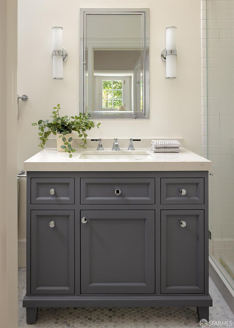 This bathroom features a dark gray vanity with a light-colored stone countertop and a rectangular undermount sink. A rectangular mirror with a metallic frame is centered above the vanity, flanked by two vertical wall-mounted light fixtures. The space is accented by a small potted plant on the counter and a stack of folded towels, set against a neutral wall and a patterned tile floor.