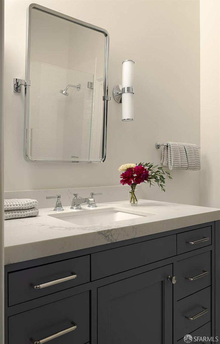 This clean and modern bathroom vanity features a dark gray cabinet base topped with a light-colored stone countertop and a rectangular undermount sink. A sleek, rectangular mirror with a metallic frame is mounted above the vanity, complemented by a contemporary wall-mounted light fixture. A small vase of fresh flowers adds a touch of color, while a towel rack with folded linens completes the polished, functional aesthetic.