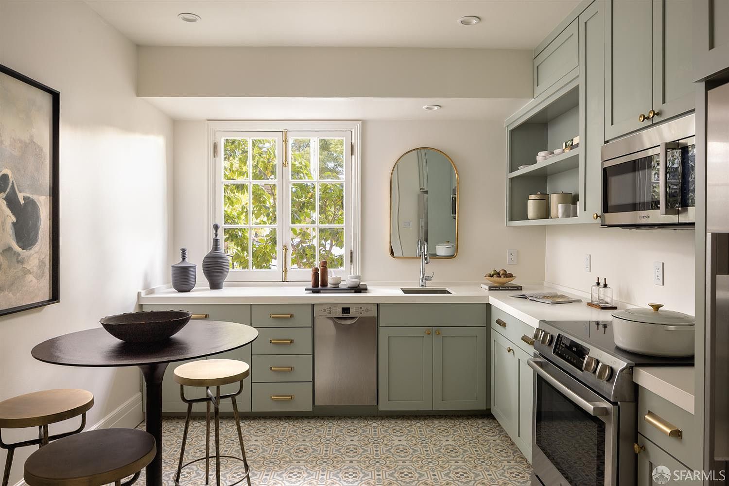 This charming, compact kitchen features soft sage-green cabinetry paired with white countertops and a distinctive patterned tile floor. A central window above the sink provides natural light, while a small round dining table with stools creates an intimate breakfast nook. The space is well-appointed with stainless steel appliances, including a dishwasher and a microwave, offering a functional and stylish design.