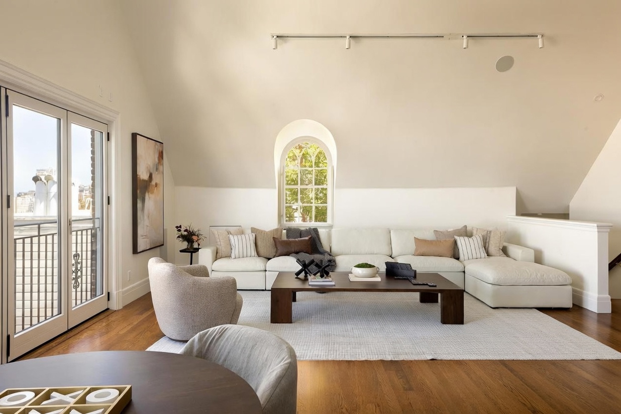 This bright and airy living room features a large, neutral-toned sectional sofa arranged around a dark wood coffee table on a textured area rug. The space is highlighted by a distinctive arched window and a set of glass doors that open onto a balcony, allowing for ample natural light. The room is decorated with a modern aesthetic, including a large piece of abstract wall art and a comfortable accent chair, creating a sophisticated and inviting atmosphere.