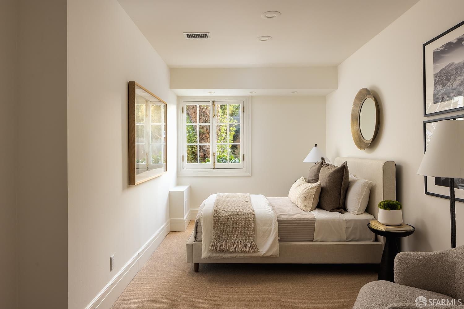 This serene bedroom features a neutral color palette with soft beige walls and matching carpeting, creating a calm and inviting atmosphere. A plush bed with layered pillows and a textured throw sits centered against the wall, flanked by a small side table with a decorative plant. The room is illuminated by recessed lighting and a bedside lamp, while a window with white muntins provides a view of greenery outside.
