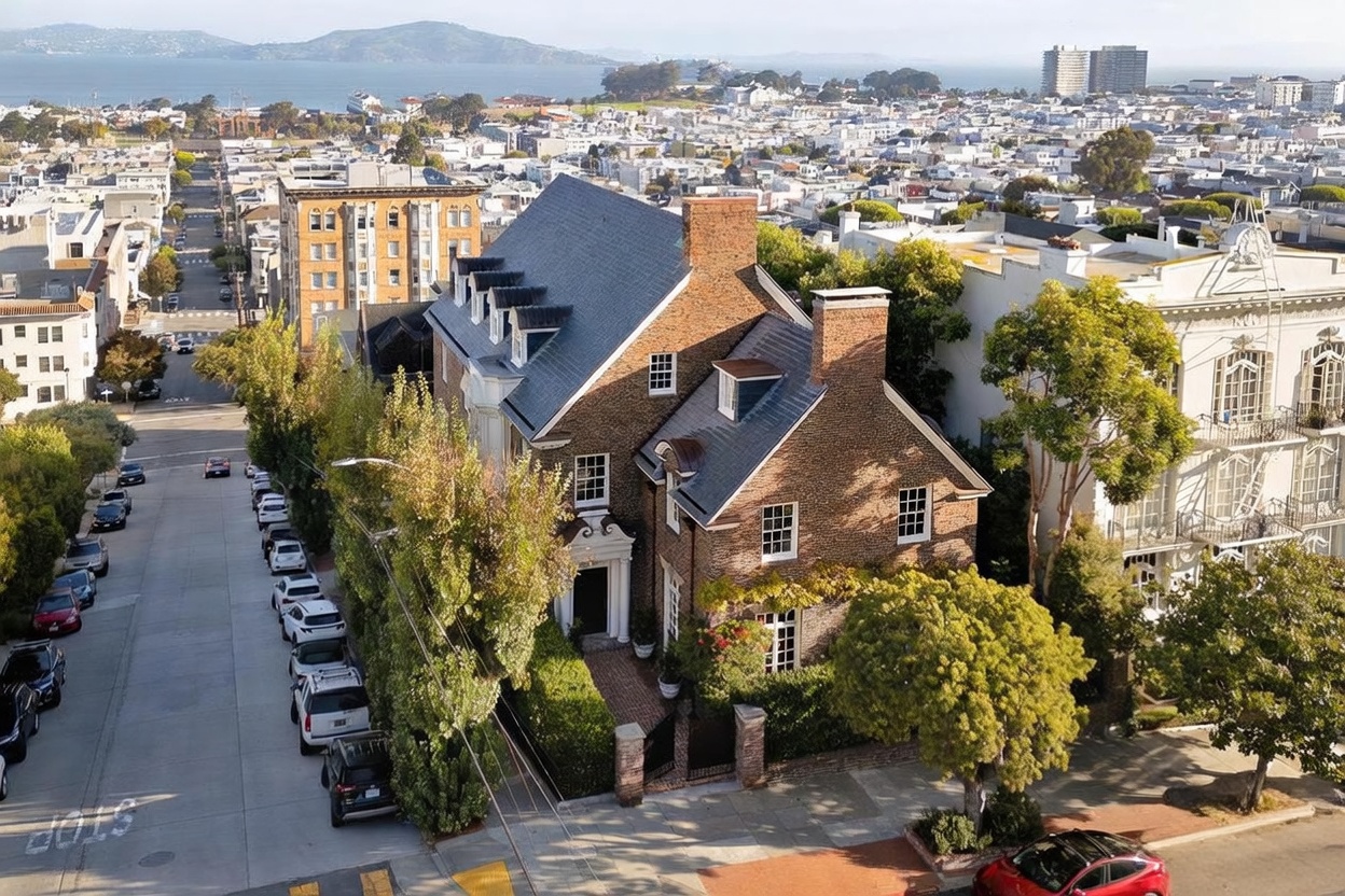 This high-angle aerial perspective captures a classic brick residence situated on a corner lot in a dense urban neighborhood. The home features a steep gabled roof, multiple dormer windows, and a charming brick-paved entryway tucked behind mature landscaping. The surrounding area showcases a mix of historic architecture and city streets, with a scenic view of the bay and distant hills in the background.