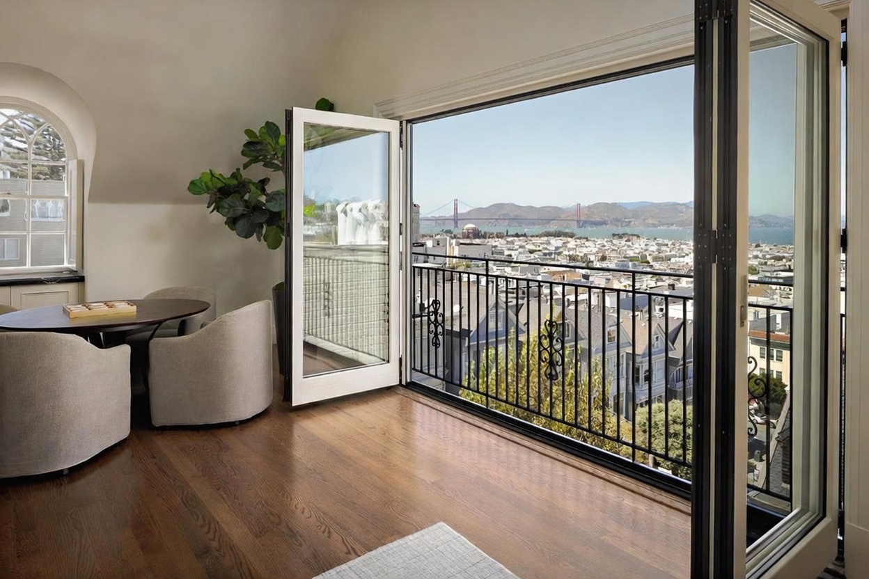 This elegant interior space features rich wood flooring and a dining area with two upholstered chairs and a dark oval table. Large, folding glass doors open onto a private balcony, providing an expansive, unobstructed view of the San Francisco cityscape and the Golden Gate Bridge. The room is bright and airy, with a large potted plant adding a touch of greenery to the sophisticated, neutral-toned decor.