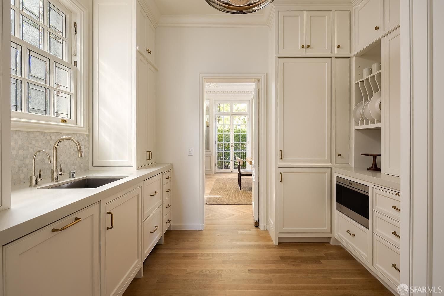 This bright and elegant butler's pantry or secondary kitchen area features crisp white cabinetry, light wood flooring, and a sleek stone countertop with an integrated sink. The space is framed by a large window with a decorative grid pattern on the left and a doorway leading to a sunlit dining area in the background. The design is clean and sophisticated, offering a seamless transition between rooms with high-end finishes and built-in storage.