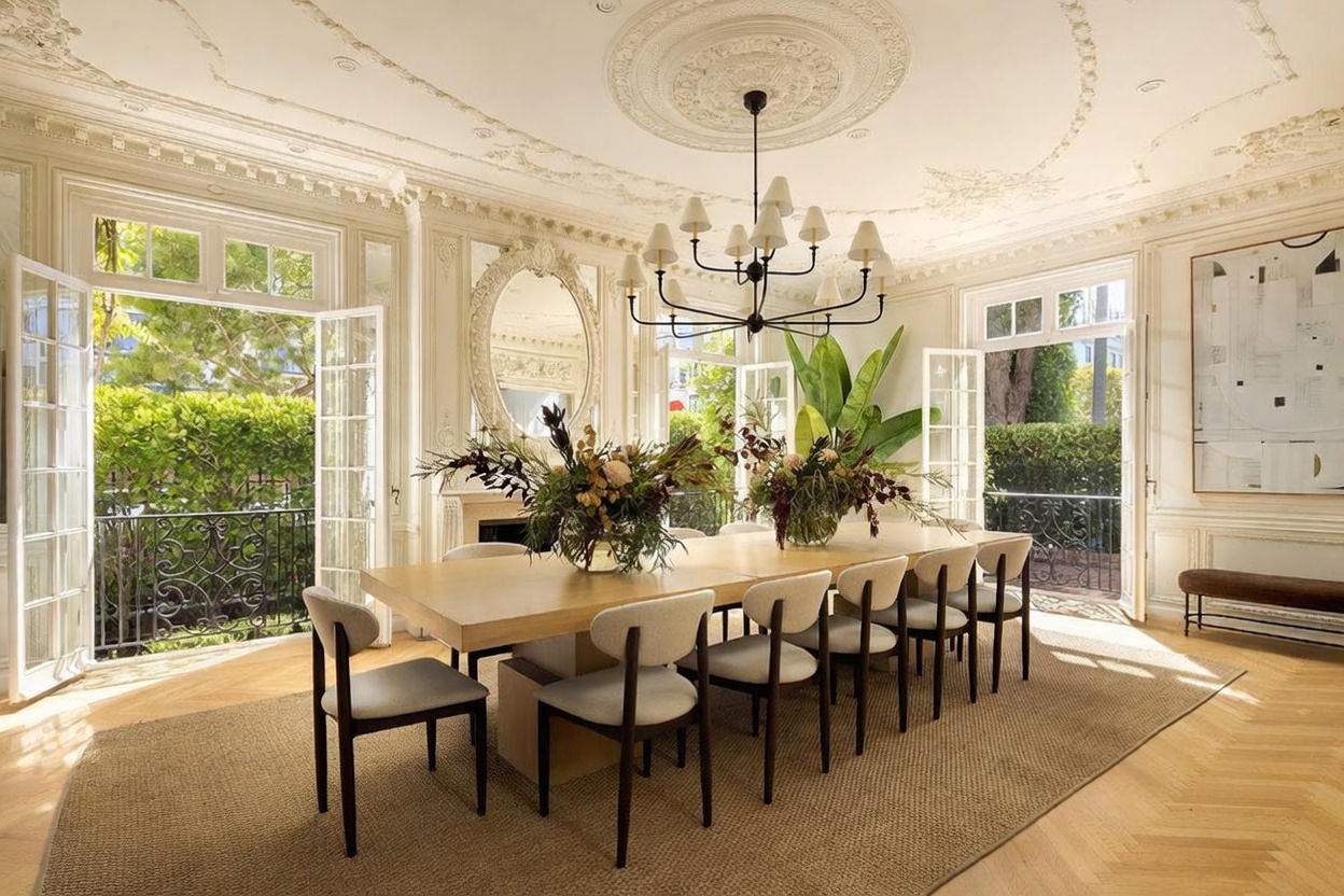 This elegant dining room features a long, light wood table surrounded by modern, upholstered chairs, set beneath a sophisticated multi-arm chandelier. The room is adorned with intricate crown molding and ceiling medallions, while large glass doors open to a lush, green outdoor space. A large, ornate mirror hangs above a fireplace, and a contemporary piece of art adds a refined touch to the bright, airy atmosphere.