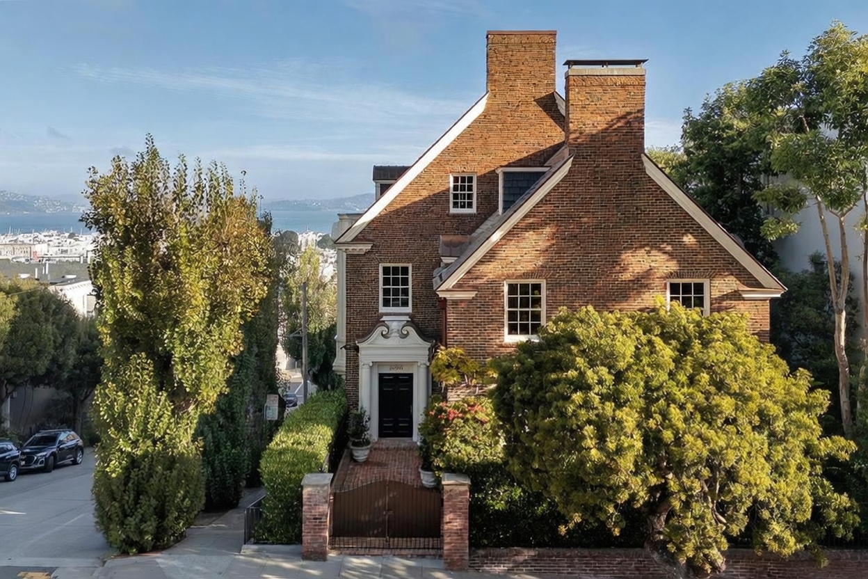 This stately brick residence features a classic architectural design with a prominent chimney and a symmetrical facade. The home is set back from the street behind a gated brick entrance and lush landscaping, offering a sense of privacy and elegance. A clear view of the bay and city skyline in the distance enhances the property's desirable location and elevated perspective.