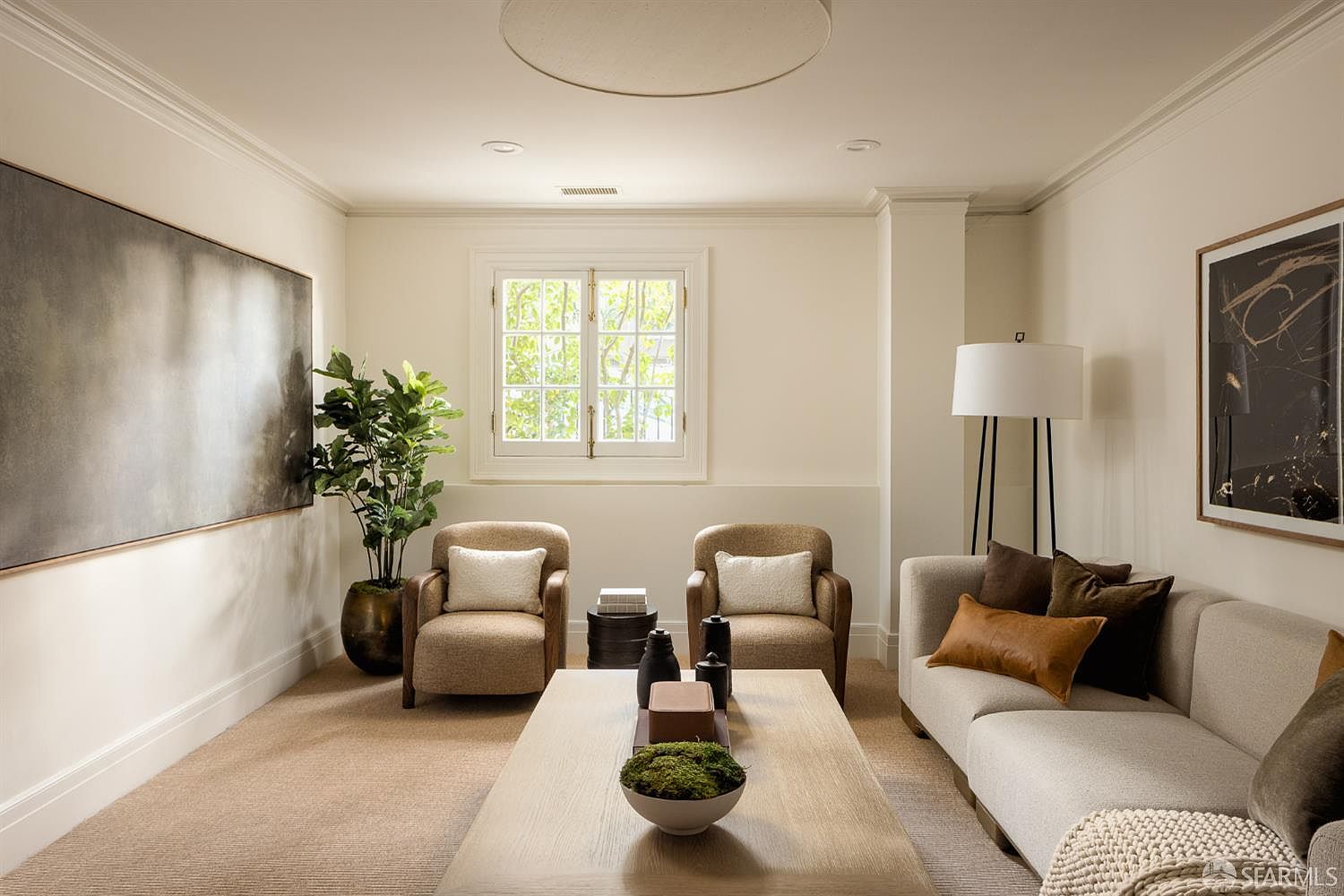 This sophisticated living room features a neutral color palette with a plush light-toned sofa, two matching armchairs, and a large wooden coffee table centered in the space. The room is accented by two large pieces of abstract wall art, a potted plant, and a tall floor lamp, creating a calm and inviting atmosphere. A central window with white trim allows natural light to fill the room, highlighting the textured carpet and clean, minimalist aesthetic.