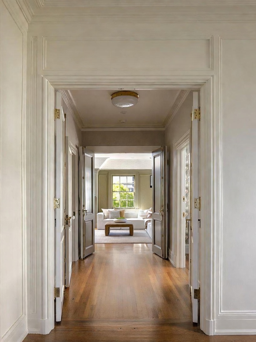 This perspective captures a bright, elegant hallway leading through a set of open double doors into a sunlit living area. The space features polished light wood flooring, crisp white trim, and classic architectural molding that frames the transition between rooms. The view through the doorway highlights a comfortable seating arrangement centered around a low wooden coffee table, creating an inviting and cohesive flow between the home's interior spaces.