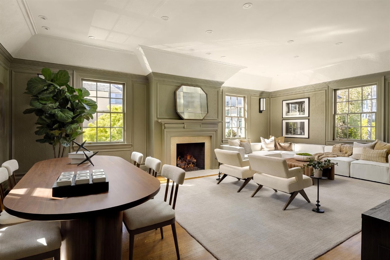 This elegant, open-concept living and dining area features sage green paneled walls and a sophisticated, neutral color palette. A central fireplace with a stone surround and a geometric mirror serves as the focal point, flanked by a large sectional sofa and comfortable armchairs. The space is bathed in natural light from multiple windows, creating an inviting and airy atmosphere perfect for entertaining.